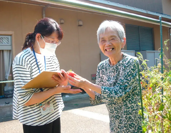 職員が利用者から笑顔で野菜（ミニトマト）を受け取っている屋外での和やかな交流の様子